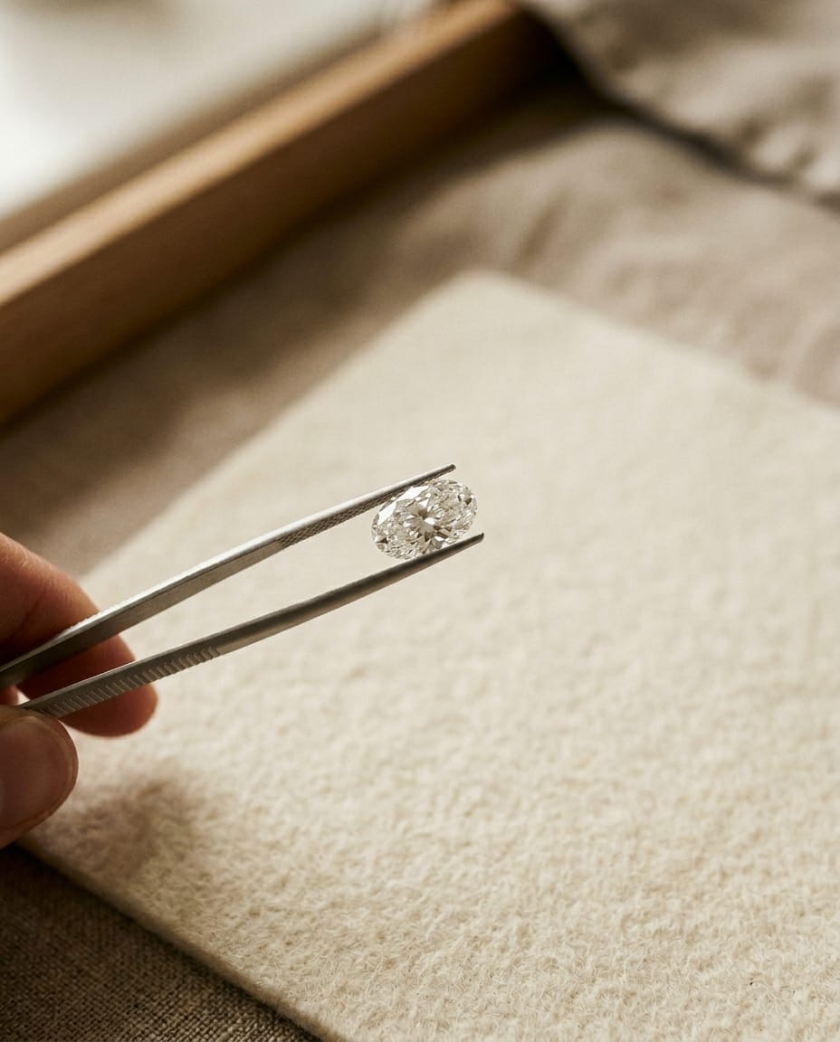 An oval diamond held in jeweller's tweezers above a soft linen tray.